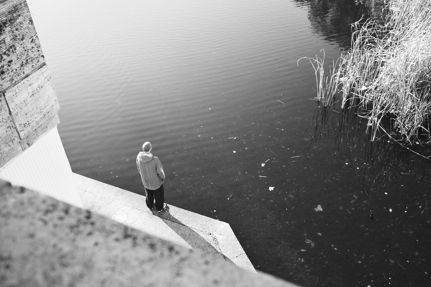 Person in grauer Jacke steht auf Betonufer und blickt auf ruhiges Wasser mit Schilfrohr in Schöneberg